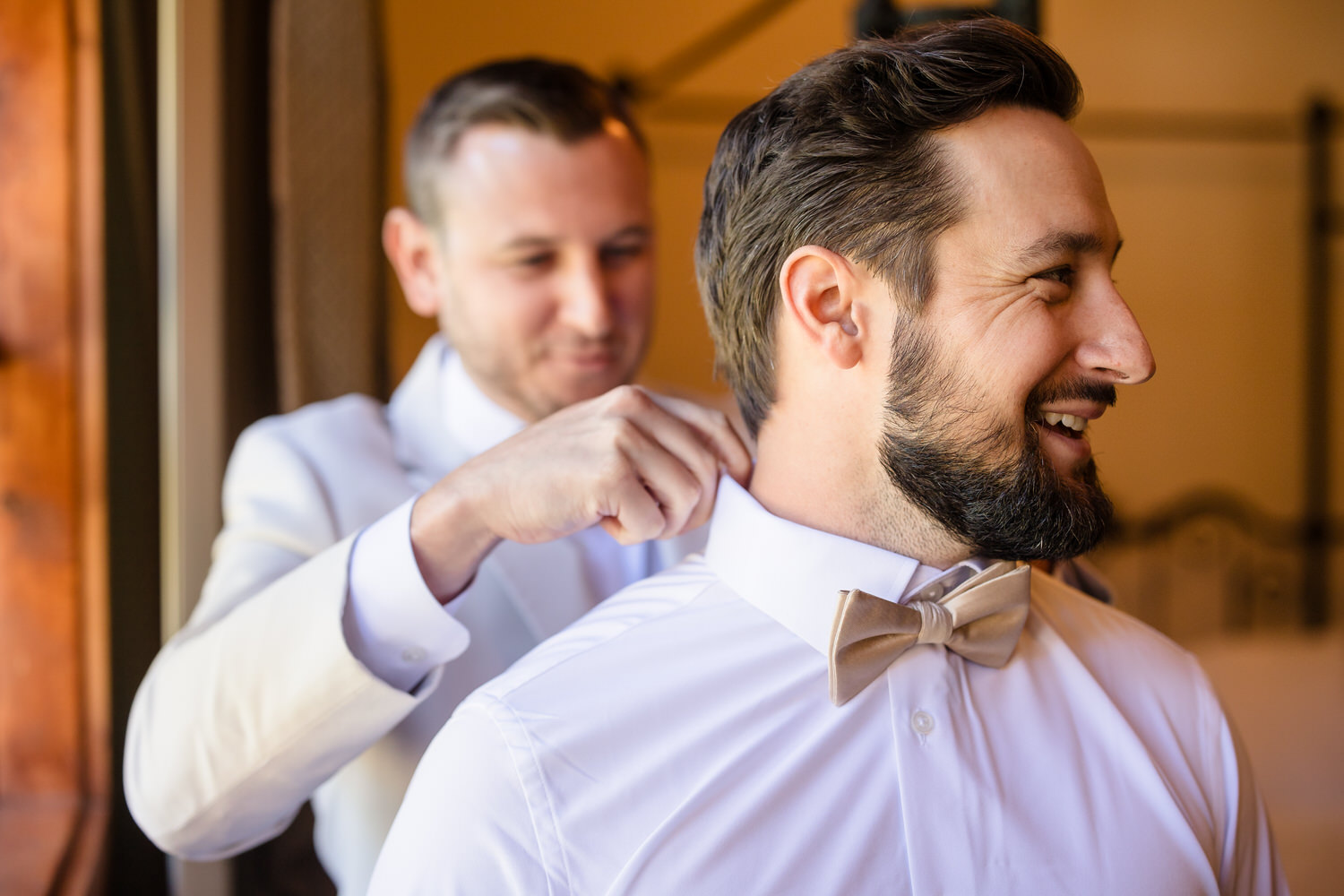 A man in a white suit adjusts the collar of a smiling man in a white dress shirt and tan bow tie.