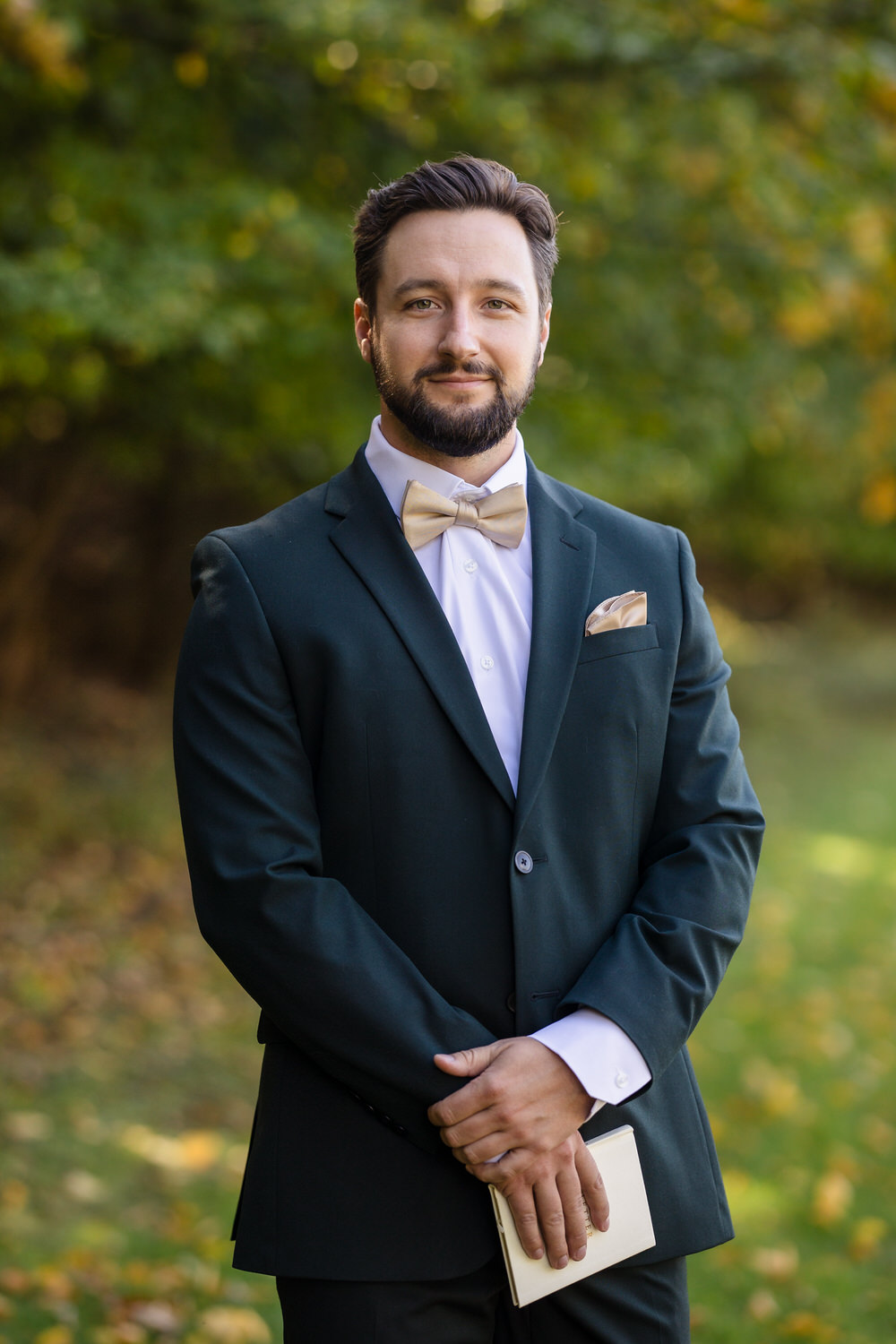 A bearded man in a dark suit, white shirt, and tan bow tie holding a small book.