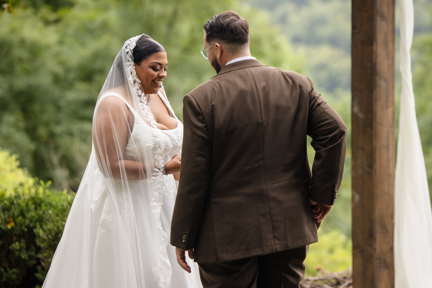 A bride in a white wedding dress and veil smiles at her groom, who stands with his back to the camera in a brown suit.
