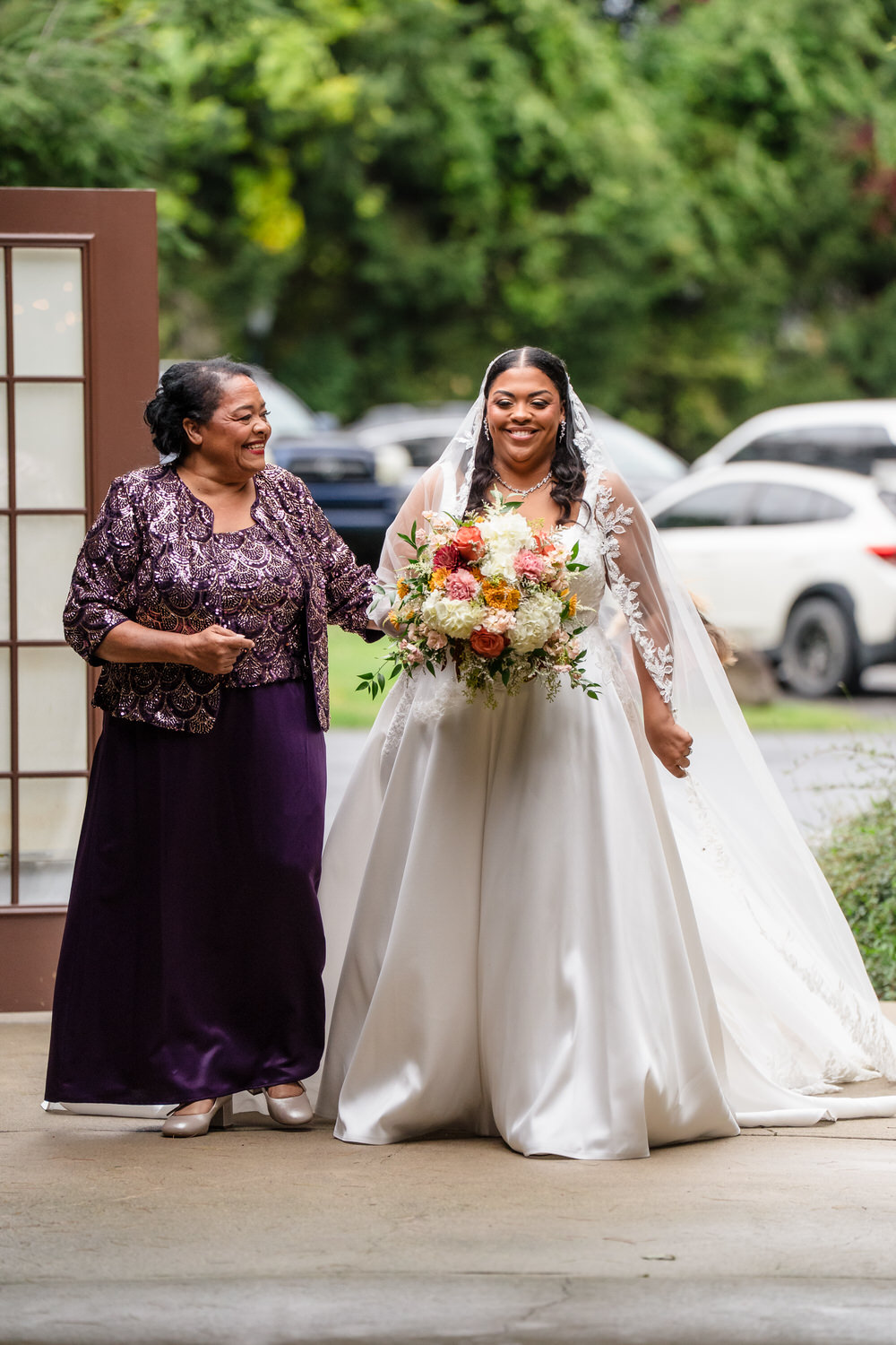 A smiling bride in a white wedding gown and veil walks arm-in-arm with an older woman in a purple dress and matching jacket, both walking outdoors.