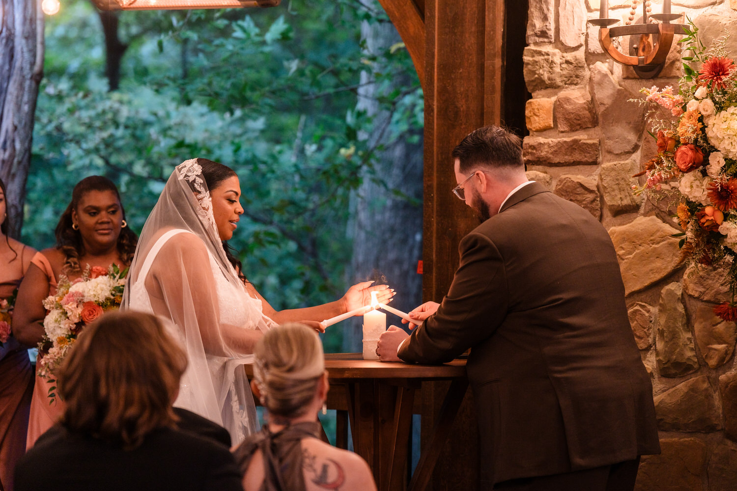 A bride and groom light a central pillar candle together during an outdoor wedding ceremony.
