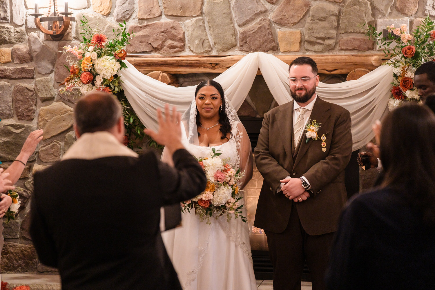 A bride and groom standing before an officiant at a wedding ceremony in front of a stone fireplace.