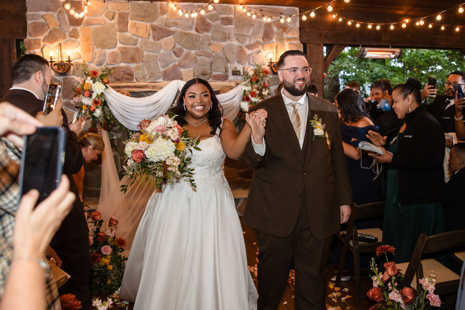 A smiling bride in a white wedding gown and a groom in a brown suit holding hands while walking down the aisle, surrounded by wedding guests.