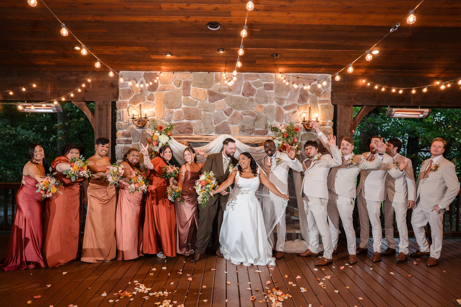 A wedding party stands on a wooden deck in front of a stone fireplace decorated with flowers and string lights, with the bride and groom at the center.