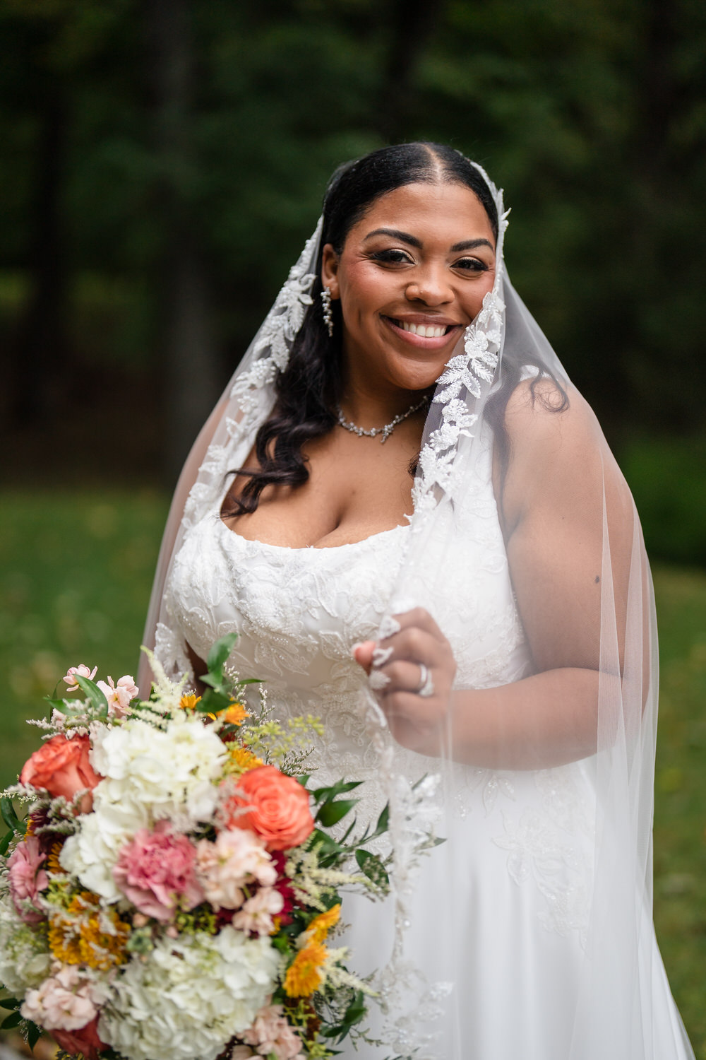 A smiling bride wearing a white wedding dress and a lace-trimmed veil holds a colorful bouquet of flowers.