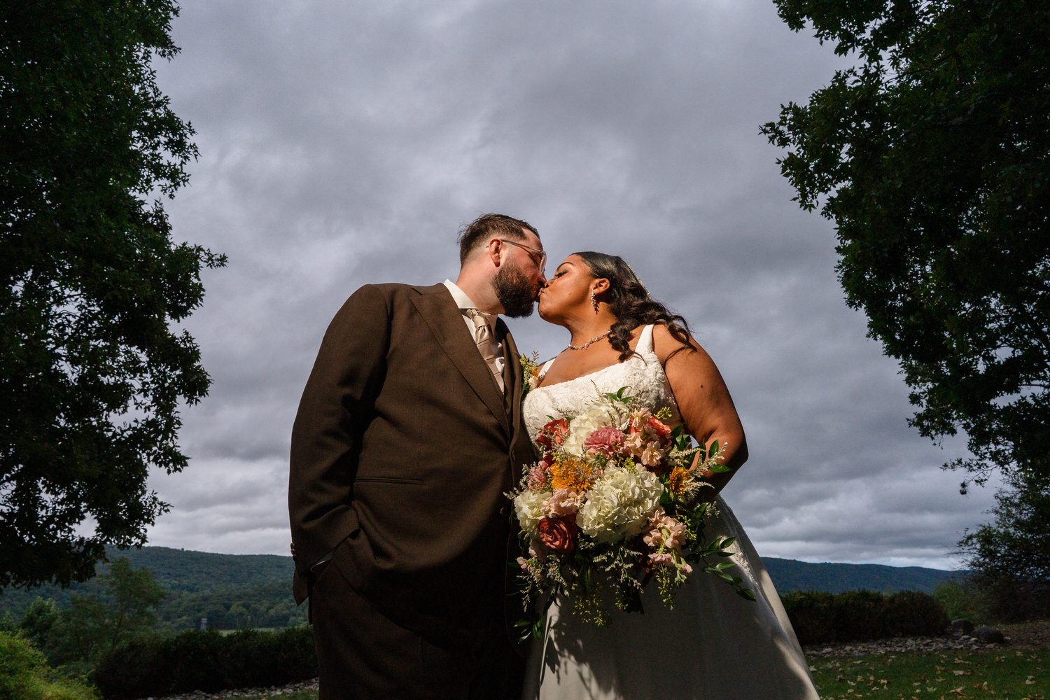A bride and groom in wedding attire kiss outdoors under a cloudy sky.