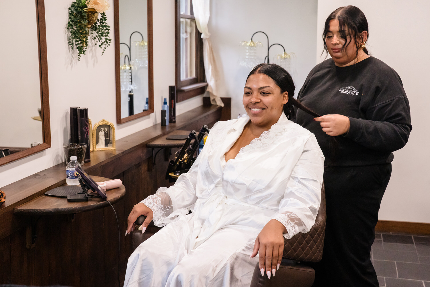 A woman in a white lace robe sits in a salon chair while a stylist brushes her hair.