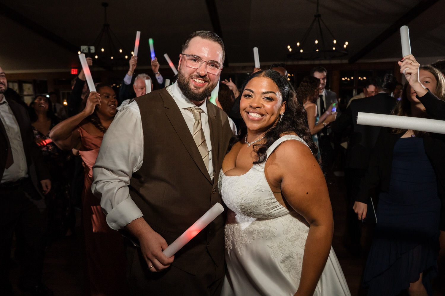 A smiling bride and groom pose together on a dance floor while holding glowing light sticks at a wedding reception.