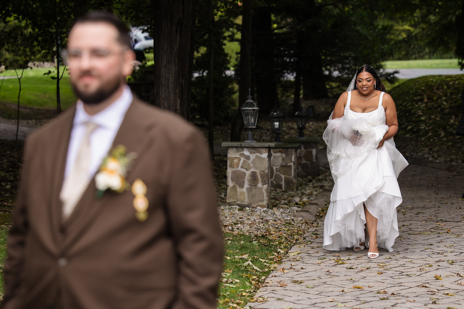 A bride in a white wedding dress walks toward a groom in a brown suit during an outdoor first look.