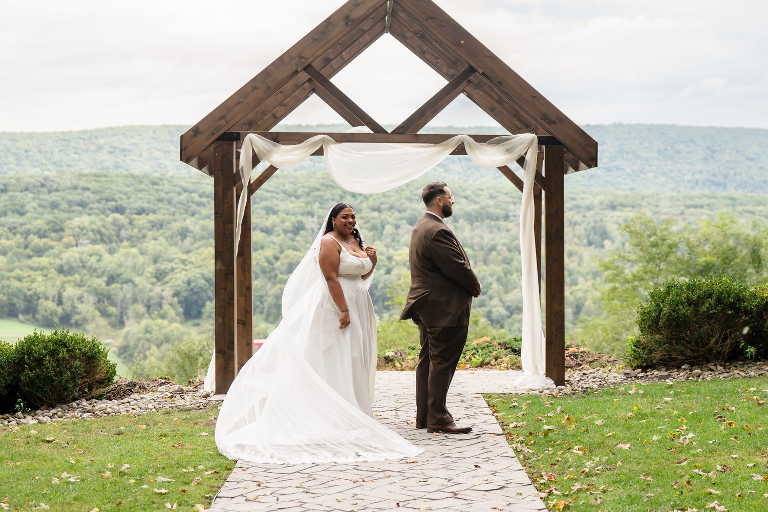 A bride in a white wedding dress with a long veil stands near a groom in a brown suit under a wooden wedding arch overlooking a mountain landscape.