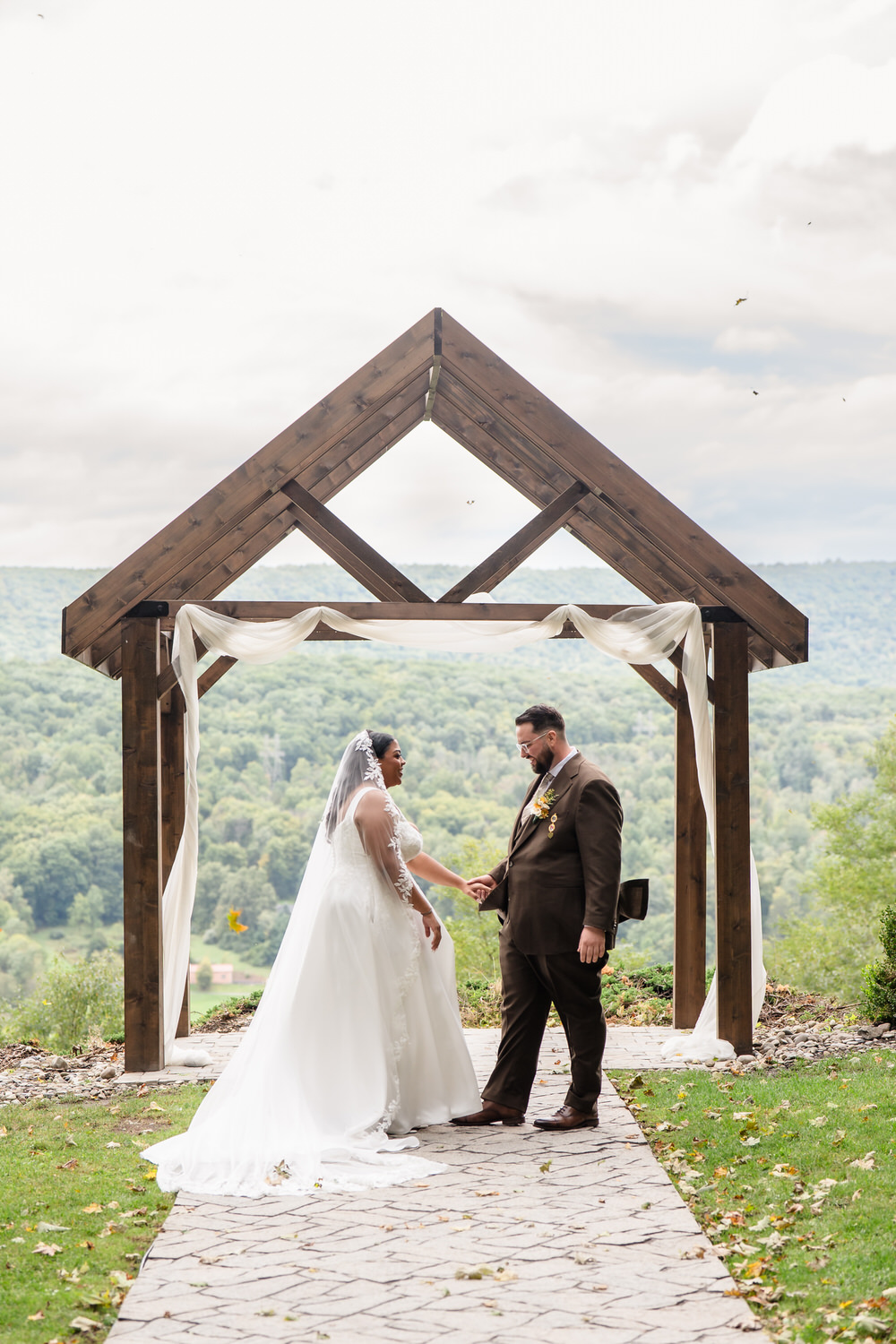 A bride in a white wedding gown and a groom in a brown suit holding hands while standing under a wooden archway with a mountain view in the background.